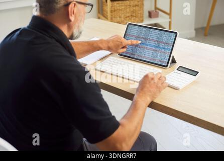 Senior man analyzing spreadsheet on tablet with calculator at tidy home office Stock Photo
