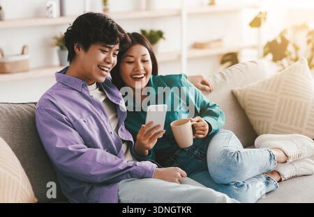 Positive young Asian couple using smartphone and drinking coffee while resting on couch at home, free space Stock Photo
