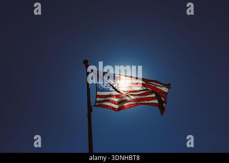 The scene shows an American flag flying on a flagpole. Sunlight shines from behind, giving the flag a bright outline. The sky is clear and blue. This Stock Photo