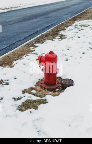 A red fire hydrant is located on the edge of a snowy field next to a dark asphalt road. Snow covers the ground, and the sun shines brightly in the sky Stock Photo