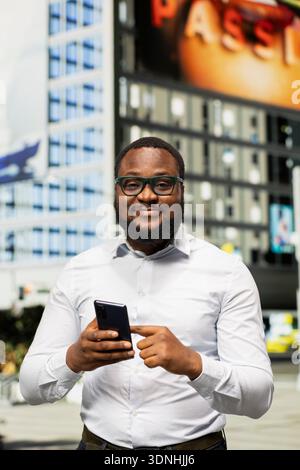 Black adult man navigating on mobile phone with map app to walk city center, admiring skyscrapers and vibrant vibe in a metropolitan area. Confident street style on urban avenue. Stock Photo
