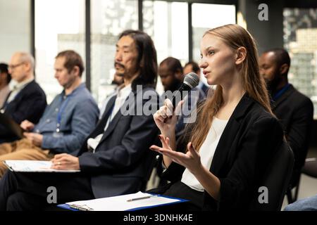 Young woman journalist reading questions from clipboard and taking notes, interacting with presenter via microphone in conference room. Writing answers for press purposes at business summit. Stock Photo