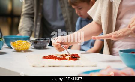 Family enjoys cooking for a cozy pizza day together at home, spreading sauce on the homemade dough for pizza. Cooking time captures happiness and bonding through delicious meal. Stock Photo