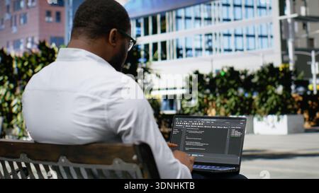 African american guy works on programming code on terminal window, sitting on a bench downtown and working on IT with his laptop in sunlight. Professional attitude in a dynamic city life. Camera B. Stock Photo