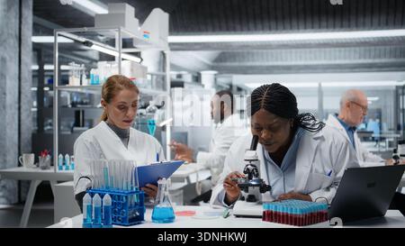 Biologist analyzes cellular samples on microscope while coworker writes research notes. Team of female scientists working together, examining sample in lab with high tech optical device gear, camera A Stock Photo