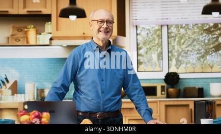 Portrait of senior man enjoying quiet moment in her home kitchen, resting near counter with coffee and pastry. Relaxed wellbeing, joyful lifestyle and calm mood of retired life indoors. Camera B. Stock Photo
