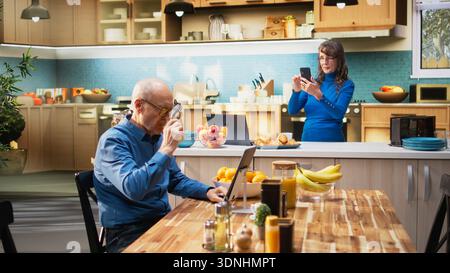 Portrait of elderly natural woman scrolling on smartphone in apartment kitchen enjoying relaxed domestic chores. Older adult reflecting joyful lifestyle and satisfied retired living. Camera A. Stock Photo