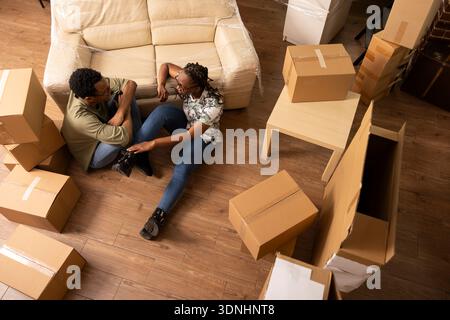 Overhead view of black couple sitting comfortably on living room floor, surrounded by moving boxes. Young spouses enjoy leisure time and conversation, resting together on relocation day in new home. Stock Photo