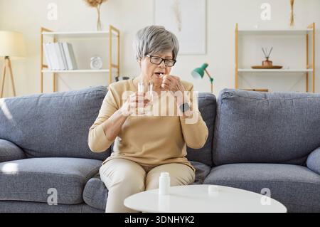 Senior woman taking pill dose at home, holding glass of water and capsule Stock Photo