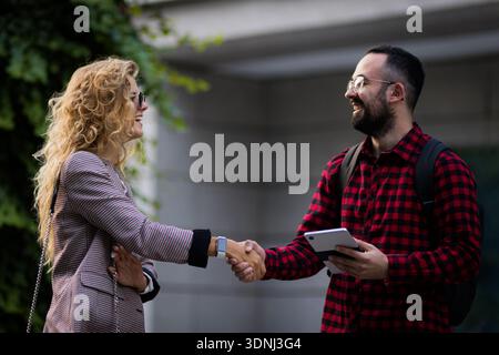 Friendly outdoor meeting between two people sharing a handshake in casual attire. A man with a tablet engages in conversation with a woman with curly Stock Photo