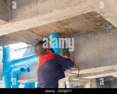 Professional plumber using an electric rotary hammer drill to create a hole in a concrete beam for securing blue PVC water pipes at a residential cons Stock Photo