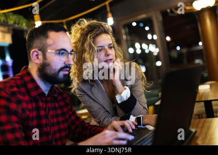 Focused colleagues collaborating on a laptop in a cozy, dimly lit coffeeshop. Young man in checkered shirt and woman in blazer intensely engaged in Stock Photo