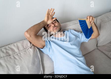 Tired man suffering from overheating and migraine at home, lying on sofa waving hand paper fan. Stock Photo