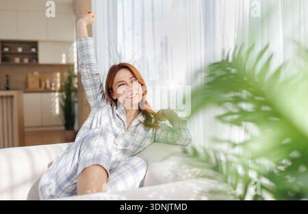 A woman in pajamas is sitting on a sofa and stretching her arms in a bright room. She seems relaxed and cheerful in the morning light. Stock Photo