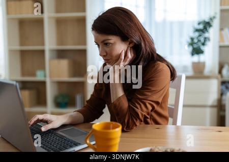A woman sits at a table using a laptop while focused on her work. A cup is beside her, and sunlight comes through the window. Stock Photo