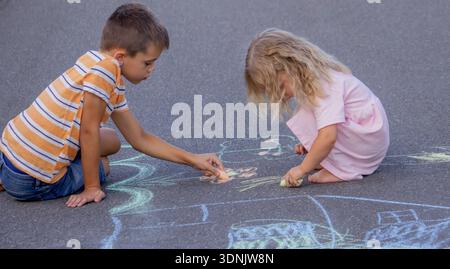 Brother and sister drawing with chalk on a path in the park on a sunny summer day. Stock Photo