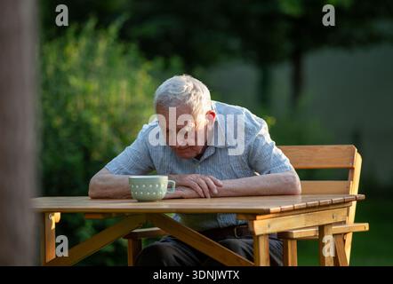 Pensioner sitting in his garden, hunched over a wooden table and looking down at coffee mug Stock Photo