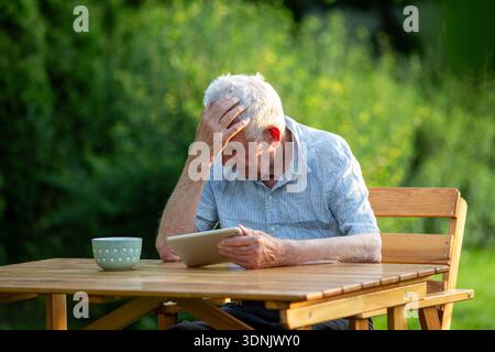 Male pensioner holding his head. He's frustrated at something on the digital tablet in his hand while sitting at wooden table in a sunny garden Stock Photo