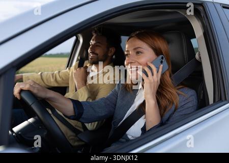 Pretty redhead woman talking on a phone with smile on her face while driving a modern car, man happily sits next to her Stock Photo