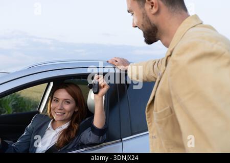 Redhead businesswoman holding up keys while sitting in car, proudly showcasing her newest purchase from the man leaning on the vehicle Stock Photo
