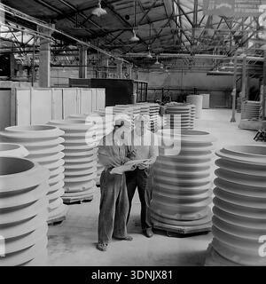 Slaviansk, Ukrainian SSR, 1960s. An archive photo of two engineers reviewing technical blueprints amidst rows of massive high-voltage ceramic insulators at the Slaviansk ZVI plant. The image highlights the scale of industrial production and quality control at this major Soviet factory in the Donetsk region. Large-scale electrical porcelain products fill the workshop, illustrating the peak of the Soviet energy industry's power. A classic example of industrial heritage, technical expertise, and the peaceful Donbas economic era. Stock Photo