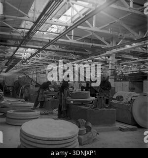 Slaviansk, Donetsk region, Ukrainian SSR, USSR. 1960s. An interior view of a bright workshop at the High-Voltage Insulator Factory (ZVI). Workers shape large flat ceramic insulator components on spinning molds using heavy mechanical levers. Stacks of raw wet porcelain discs fill the foreground under a complex lattice roof structure. This archive image captures the primary clay forming stage of mass electrical manufacturing. A nostalgic historical view of heavy industry and the daily labor routine of the peaceful Donbas before the war. Stock Photo