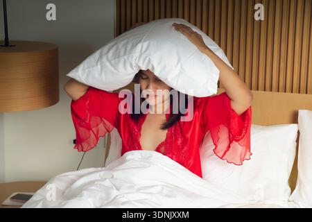 Woman in red satin nightgown and robe covering her head with a pillow while lying in bed. Bright light causes irritation and insomnia, expressing slee Stock Photo
