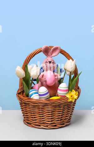 Multicolored Easter eggs in a basket with spring flowers: white tulips and yellow daffodils and pink Easter knitted bunny with pink egg on gray table. Easter concept Stock Photo
