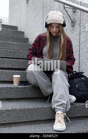 Woman in her 20s sitting on concrete steps at campus, using laptop wearing beanie and headphones Stock Photo
