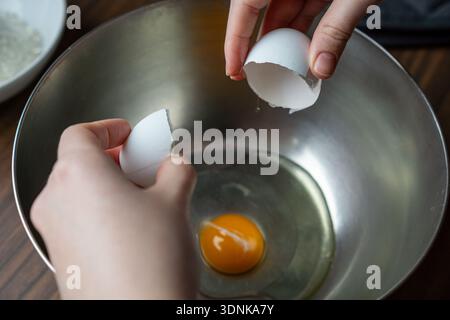 Close-up of empty egg shells held over a metal mixing bowl with yolk and egg white inside. Stock Photo