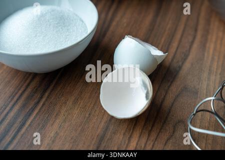 Close-up of empty egg shells lying on a wooden kitchen countertop next to a bowl of sugar and a metal whisk. Stock Photo