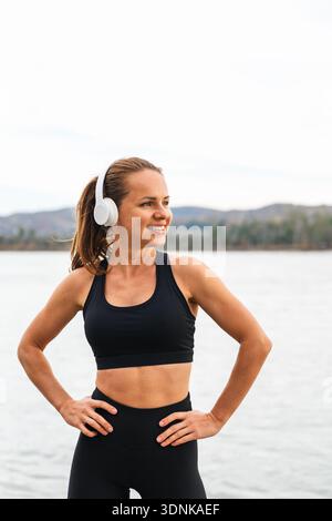 Smiling sporty female resting after workout and enjoying music in nature with river on background. Stock Photo