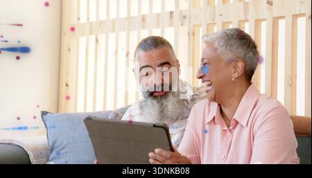 Sharing mature couple in striped and pink shirts leaning on sofa at home, man holding tablet Stock Photo