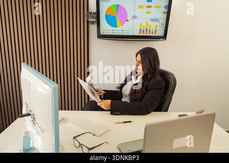 Businesswoman reviewing financial charts and reports while working in an office with a screen showing analytics Stock Photo