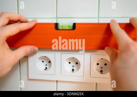 Technician's hands carefully positioning an orange spirit level above a new strip of white electrical outlets, verifying the even placement during a construction project Stock Photo