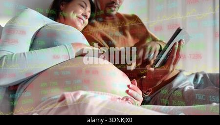 Reclining pregnant woman showing belly in white top man holding tablet on sofa with stock tickers Stock Photo