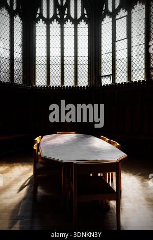 Simple wooden table with chairs stands in front of tall leaded windows in a dark historic interior. Stock Photo