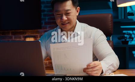 Smiling middle-aged Korean business man typing laptop review papers corporate good financial results in night office Chinese businessman smile Stock Photo