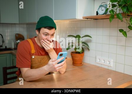Young man receiving shocking news on smartphone, covering mouth in disbelief, reacting to job cuts, economic crisis, financial problems, and unemployment issues Stock Photo