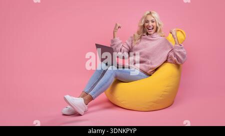 Smiling Woman in Pink Hoodie Sits on Yellow Bean Bag Chair Using Laptop Against Pink Background Stock Photo
