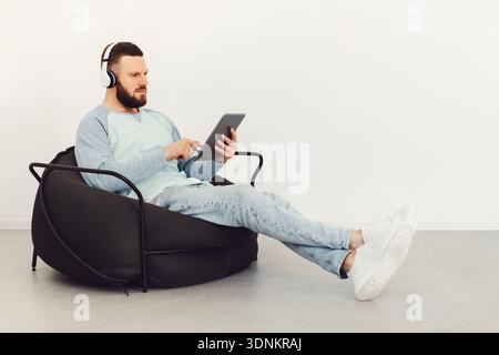 Serious Guy Using Tablet Wearing Earphones Sitting Indoor, Side View Stock Photo
