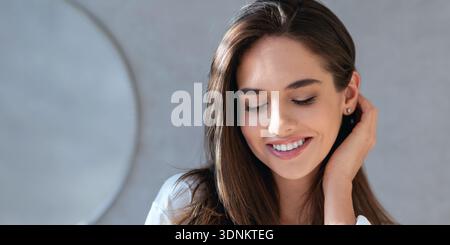 Woman Smiles While Touching Her Hair and Looking Down in a Bright Indoor Setting During Day Stock Photo
