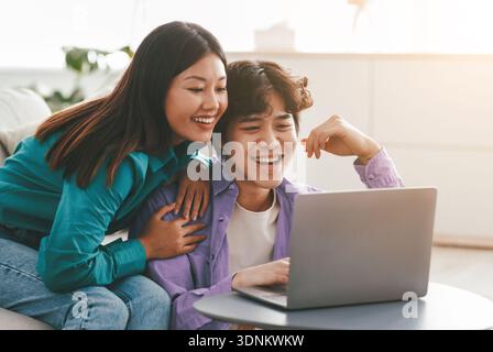 Millennial Asian couple in casual wear using laptop, having online meeting, watching interesting movie in living room Stock Photo