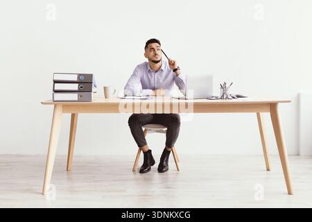 Man Sits at Desk Thinking While Surrounded by Office Supplies in Bright Room Stock Photo