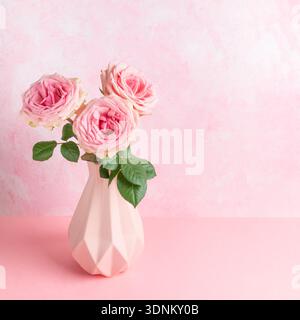 Three pink roses with green leaves are arranged in geometric vase on pink surface, complemented by textured pastel pink background, creating harmoniou Stock Photo