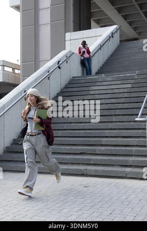 Diverse students running across urban plaza carrying notebooks and folder, one using smartphone Stock Photo