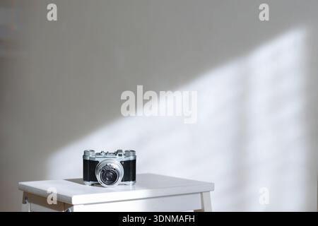 Vintage film camera placed on white stool against light wall with natural sunlight and copy space. creative hobby background. Stock Photo