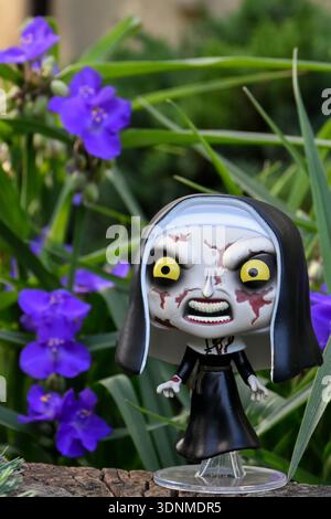 Funko Pop action figure of the Nun from The Conjuring horror movie franchise. Creepy gothic doll in garden among blue flowers on sunny day. Stock Photo