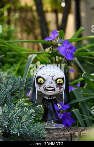 Funko Pop action figure of the Nun from The Conjuring horror movie franchise. Creepy gothic doll in garden among blue flowers on sunny day. Stock Photo