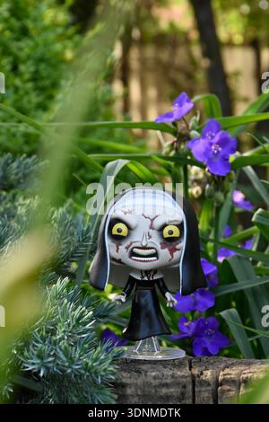 Funko Pop action figure of the Nun from The Conjuring horror movie franchise. Creepy gothic doll in garden among blue flowers on sunny day. Stock Photo